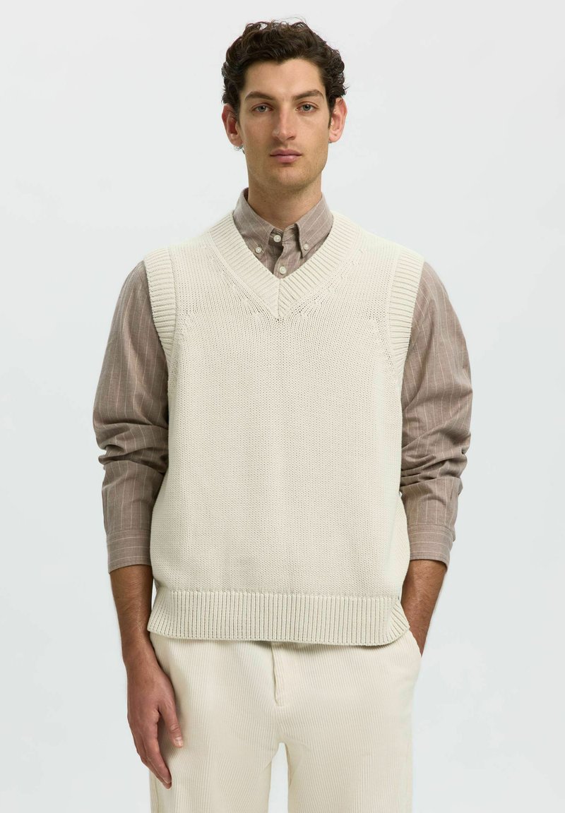 Homme portant un gilet en maille beige sur une chemise marron clair boutonnée avec un pantalon couleur crème sur un fond uni.