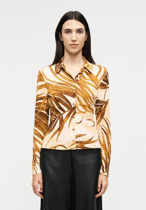 Femme aux cheveux longs et foncés portant une chemise boutonnée à manches longues avec un motif de feuilles marron et beige, et un pantalon noir, debout face à l'avant.