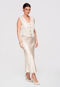 Femme portant un gilet sans manches crème avec des boutons dorés, une jupe en satin assortie, des talons à brides cheville, des boucles d'oreilles dorées et un bracelet, se tenant devant un fond blanc.