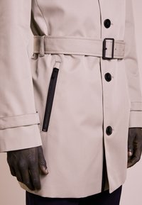 Cappotto beige con vita cinturata, dotato di bottoni neri e tasca con zip. Texture liscia, design strutturato e maniche lunghe.