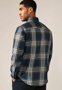 Camisa de manga larga en azul oscuro con un gran estampado de cuadros en marrón, blanco y azul claro. Presenta un cuello abotonado y puños.