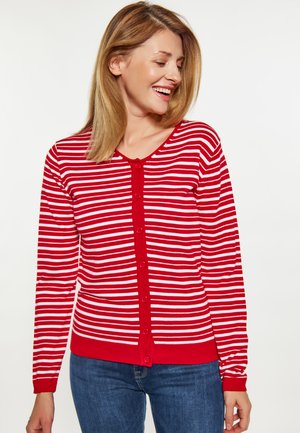 Vrouw glimlachend, draagt een rood-wit gestreepte gebuttonede cardigan en blauwe jeans, kijkt naar links tegen een effen witte achtergrond.