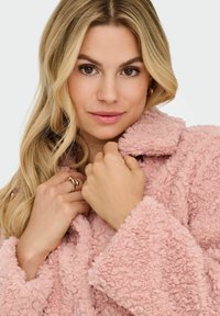 Pink fleece coat with a textured, fluffy fabric, featuring a notched collar and oversized sleeves, worn by a person holding the collar.