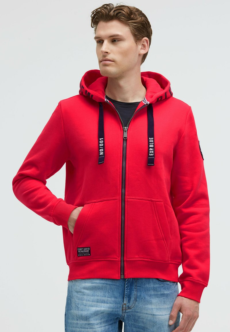 Czerwony rozpinany hoodie z kieszenią kangurzą, czarnymi sznurkami akcentującymi oraz naszywką z logo na dolnej części przodu. Wykonany z miękkiego, teksturowanego materiału bawełnianego.