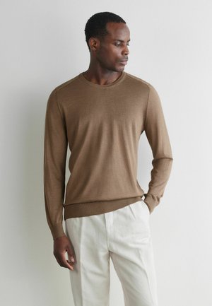 Mand i brun langærmet sweater og hvide bukser, står med den ene hånd i lommen og kigger til siden mod en ensfarvet hvid væg.