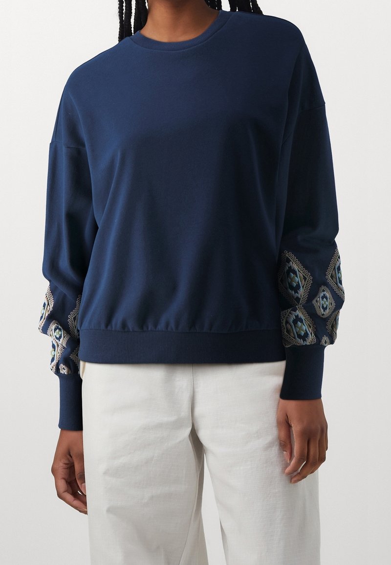 Sweat-shirt à manches longues bleu marine avec broderie en forme de losanges motifs sur les manches, porté avec un pantalon blanc.