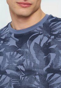 Κοντινό πλάνο ενός άνδρα που φορά μπλε μπλουζάκι με στρογγυλή λαιμόκοψη και σκούρο μπλε τροπικό φύλλο μοτίβο. Ορατά είναι μόνο το κάτω μέρος του προσώπου και το πάνω μέρος του στήθους.