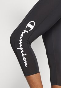 Czarne legginsy capri o gładkiej fakturze, z białym logo "Champion" nadrukowanym pionowo wzdłuż zewnętrznej strony.
