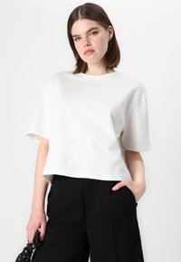 Tricou scurt alb din bumbac, cu un design texturat și detalii de cusătură subtile. Modelul poartă pantaloni largi negri.