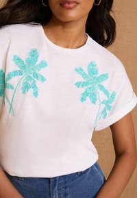 Balta īsa piedurkne T-kreklu ar turkoza izšūtām palmu lapām. Mīksta tekstūra ar sarullētām piedurknēm, kombinēta ar zilām džinsa biksēm.