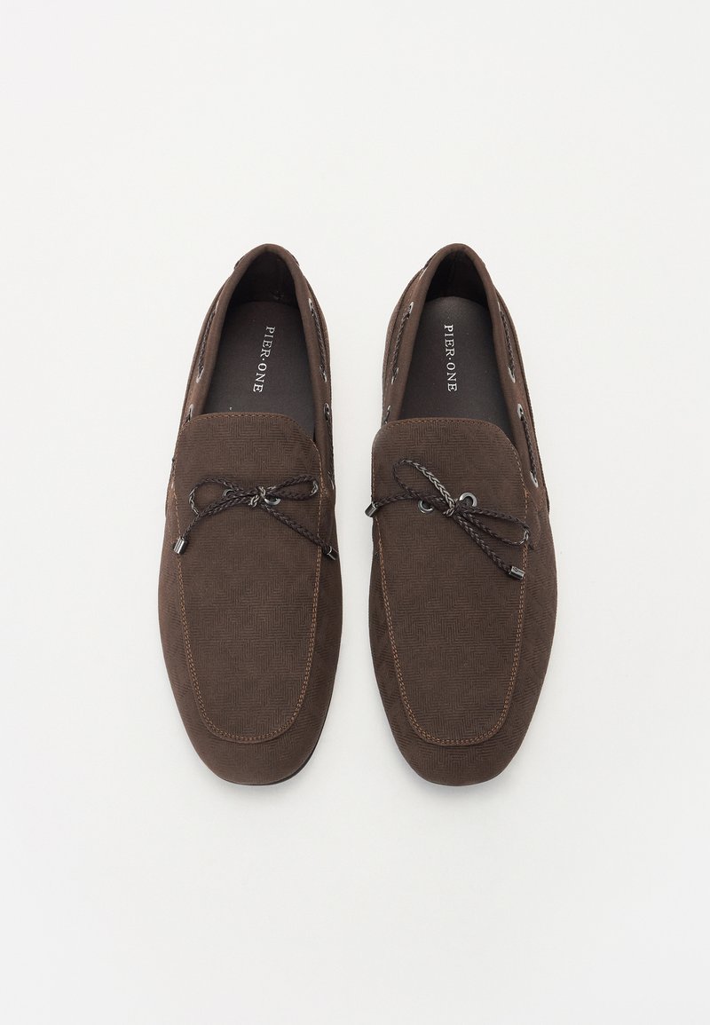 Zapatos marrones sin cordones con una tela texturizada, que cuentan con una línea de costura contrastante y cordones decorativos. El interior es negro con marca.