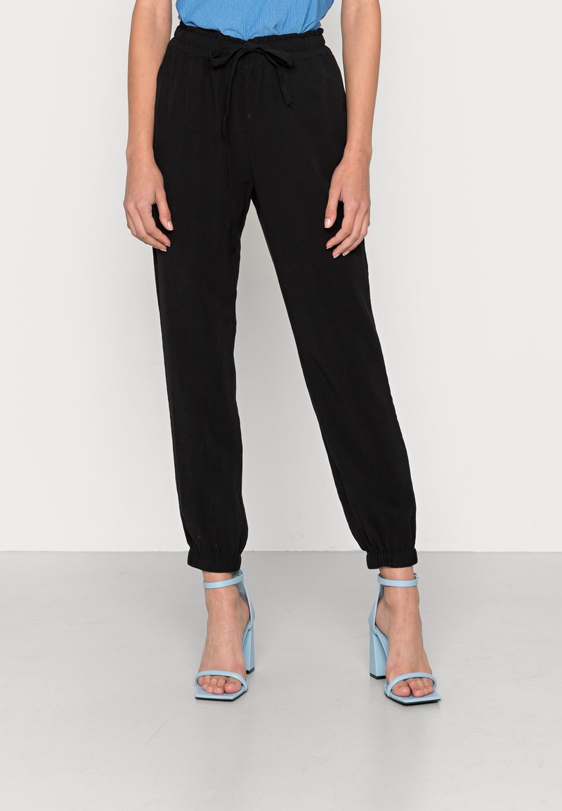 Femme portant un pantalon de jogging noir à cordon et des sandales à talons ouverts bleu clair, debout devant un fond clair uni.