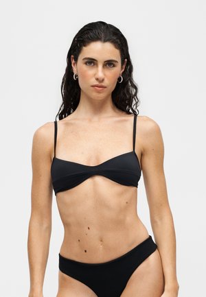 Žena s dlhými tmavými vlasmi nosí čierne bikiny s kríženým vrchným dielom vpredu a jednofarebným spodným dielom na jednotnom svetlom pozadí.