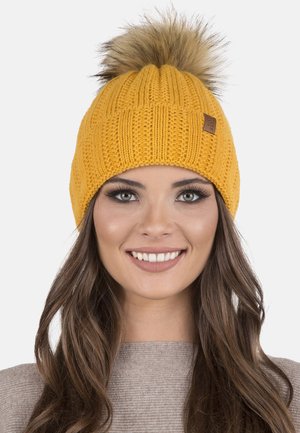 Bonnet en tricot jaune avec une texture côtelée et un pompon en fausse fourrure sur le dessus. Présente une petite étiquette en cuir sur le côté.