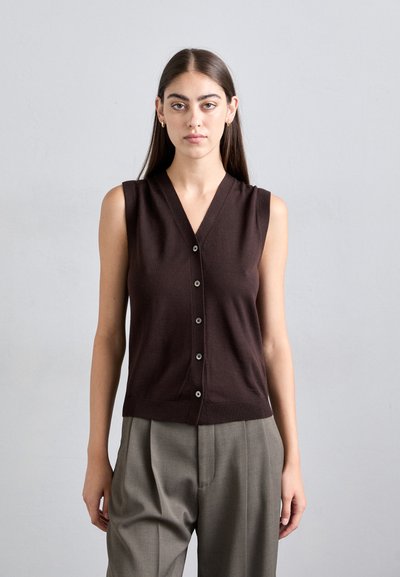 Mujer con pelo largo y oscuro que lleva un chaleco sin mangas de color marrón oscuro con botones y pantalones grises sueltos, frente a un fondo claro y liso.