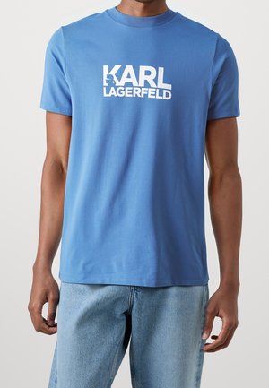 Homme portant un t-shirt bleu Karl Lagerfeld avec un logo blanc et un jean bleu clair sur un fond uni.