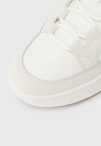 Primer plano de la punta de una zapatilla blanca con ribete de ante, cordones blancos y costuras detalladas sobre un fondo claro.