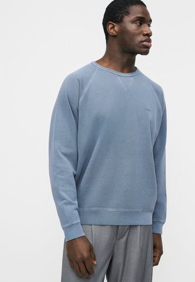 Homme portant un sweat-shirt bleu clair à col rond et un pantalon gris, faisant face à droite sur un fond blanc uni.