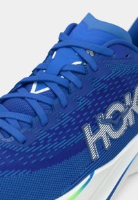 Κοντινό πλάνο ενός μπλε αθλητικού παπουτσιού με υφασμάτινη υφή, μπλε κορδόνια και λευκή σόλα με πράσινες και μπλε λεπτομέρειες.