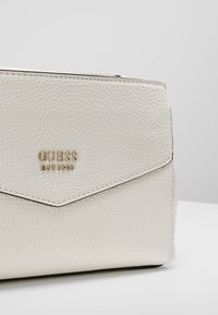 Clutch in pelle bianca testurizzata di Guess con logo dorato "GUESS EST 1981" sulla pattina anteriore su sfondo grigio.