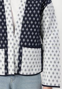 Quilted jacket with navy and white patterned fabric. Features a V-neck, long sleeves, and two front pockets. Accented with contrasting trim.