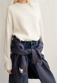 Pull en tricot crème avec un col rond, associé à un jean bleu. Une écharpe bleu marine foncé est nouée autour de la taille, ornée de boutons marron.