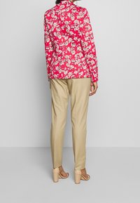 Blusa rossa con stampa floreale, a maniche lunghe, colletto e una silhouette aderente, abbinata a pantaloni beige sartoriali e sandali con tacco chiaro.