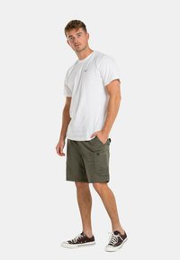 Camiseta blanca de algodón con mangas cortas y logo, combinada con pantalones cortos cargo en color verde oliva con bolsillos laterales, usados con zapatillas marrones.