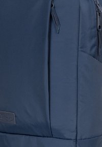 Zaino blu navy realizzato in tessuto resistente, dotato di una tasca con zip e una superficie testurizzata. Etichetta del logo visibile sulla parte frontale.