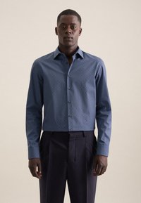 Camicia button-up blu con motivo, colletto strutturato, maniche lunghe e vestibilità slim. Indossata con pantaloni scuri e sartoriali.