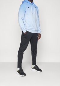 Felpa con cappuccio blu sfumata, con lacci e tasca frontale, abbinata a pantaloni sportivi neri con un piccolo logo bianco e scarpe da ginnastica nere.