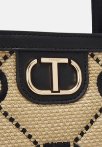 Sac à main en toile beige avec des accents noirs, présentant des éléments en métal doré et un fermoir avec logo. Des coutures décoratives noires ajoutent de la texture.