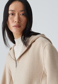 Beige Kapuzensweatshirt aus weichem Stoff, mit Kapuze und Reißverschluss. Darunter wird ein hellfarbener Pullover mit gerippter Textur getragen.