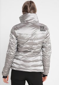 Silver puffersoft med hög krage, kviltad textur och broderad logotyp på ryggen. Den har ett glänsande, slätt tyg.