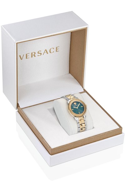 Versace kell valges karbis. Omab kuld- ja hõbemetallist rihmu, rohelise kellapea ning geomeetrilise äärise koos logoga. Pehme valge padja väljapanek.