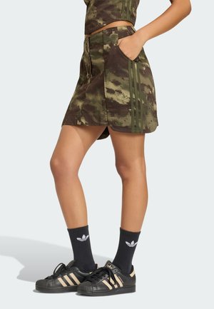 Kvinde iført et nederdel med camouflage-mønster med lommer, sorte Adidas sneakers med beige striber og sorte Adidas-logo sokker, stående sideværts.