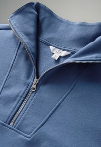 Blauwe half-rits trui van een zachte, gestructureerde stof. Voorzien van een geribde kraag en een zilveren rits, met een label dat maat M aangeeft.