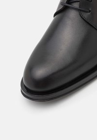 Chaussure habillée en cuir noir avec un bout arrondi, une texture lisse et des détails cousus au niveau des lacets. Présente une finition élégante et polie.