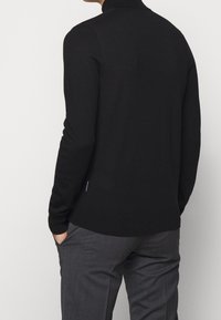 Maglione nero aderente con colletto alto, maniche lunghe e una texture liscia, abbinato a pantaloni grigi. Design minimalista senza motivi visibili.