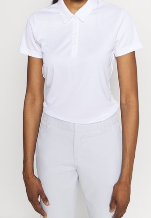 Persoon draagt een effen witte poloshirt met korte mouwen en lichtgekleurde aansluitende broek, staand tegen een neutrale achtergrond.