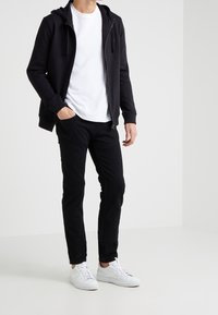 Man som bär en svart zip-up hoodie, vit t-shirt, svarta byxor och vita sneakers står mot en enkel vit bakgrund.