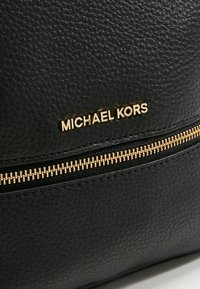 Musträne musta nahast kott, millel on kuldne tõmblukk ja bränding, mis sisaldab kuldset kirja "MICHAEL KORS."