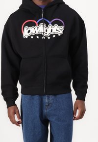 Sweat à capuche noir zippé avec un grand logo blanc et des anneaux multicolores. Comprend une poche kangourou et des manches côtelées, assorti à un jean bleu.