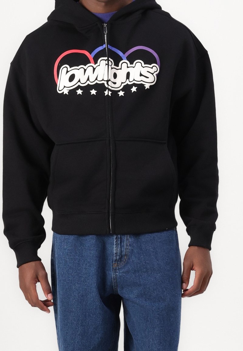 Sweat à capuche noir zippé avec un grand logo blanc et des anneaux multicolores. Comprend une poche kangourou et des manches côtelées, assorti à un jean bleu.