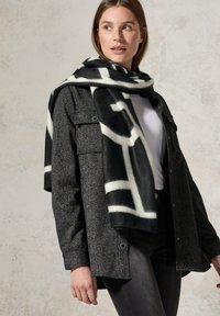 Ein schwarz-weiß gemusterter Schal aus weichem Stoff fällt über ein graues Tweed-Hemd. Das Outfit hat einen klassischen Kragen und Taschen.