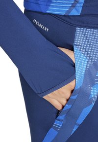 Marine Sportbekleidung aus strukturiertem blauen Stoff, mit einer Tasche und dem "AEROREADY"-Logo. Nahaufnahme einer Hand, die in die Tasche gesteckt ist.
