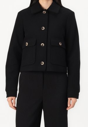 Veste noire boutonnée avec deux poches à rabat sur le devant, portée avec un pantalon noir assorti sur une personne aux longs cheveux noirs.