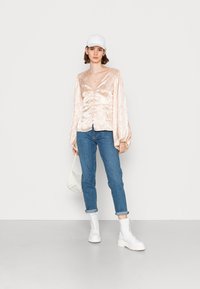 Camisa de seda blush com botões frontais e mangas bufantes, combinada com jeans azul escuro e botins brancos com atacadores, acessorizada com uma bolsa branca.