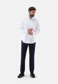 Weißes Langarmhemd mit Button-Down-Kragen, einer Brusttasche; kombiniert mit dunkelblauen maßgeschneiderten Hosen und schwarzen Schuhen.