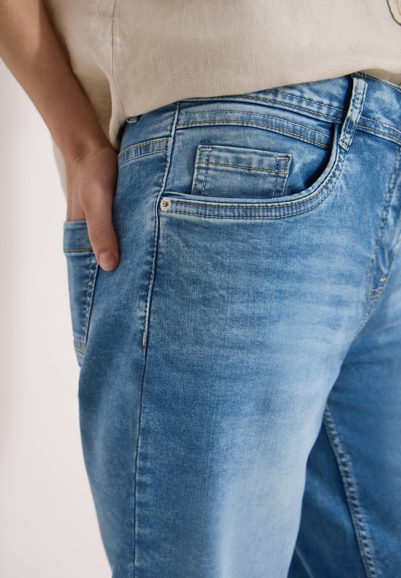 Gros plan d'une personne portant un jean en denim bleu clair avec la main placée dans la poche arrière, haut beige partiellement visible.